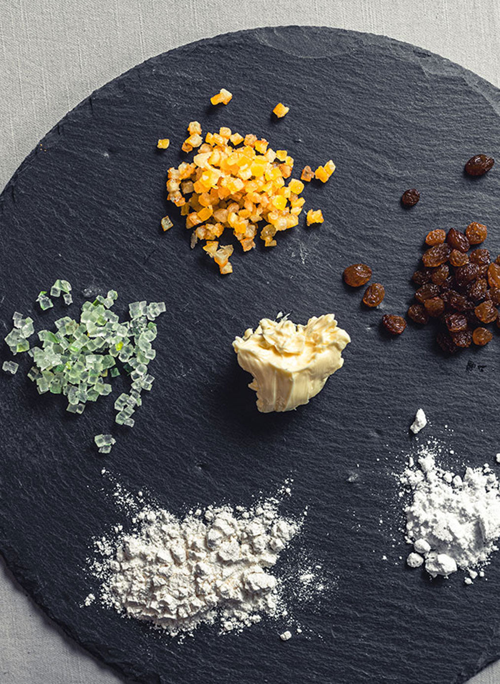 Steinernes Schneidebrett schwarz mit verschiedenen Backzutaten wir Zucker, Mehl, Rosinen, Butter usw